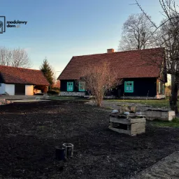 Czarny, parterowy dom z czerwoną dachówką i jaskrawozielonymi okiennicami, obok garaż z samochodem, na pierwszym planie przygotowana ziemia pod uprawy.