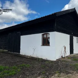 Budynek z czarną drewnianą elewacją górnej części i białą, otynkowaną dolną częścią, z małymi oknami i drzwiami, otoczony świeżo przekopaną ziemią i zieloną trawą, pod błękitnym niebem.