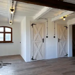 Wnętrze domu z dwiema drewnianymi drzwiami w stylu rustykalnym, białymi ścianami, drewnianym oknem i podłogą z płytek imitujących drewno, oświetlone wiszącymi żarówkami.