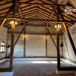 Surowe drewniane belki stropowe i kolumny w przestronnym, jasnym wnętrzu z drewnianą podłogą i oknami w drewnianych ramach. Ściany wykończone w stylu rustykalnym, widoczne lampy wiszące w stylu...