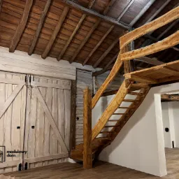 Wnętrze domu z drewnianą konstrukcją dachu, antresolą i schodami z surowego drewna oraz stylizowanymi na stare, drewnianymi drzwiami stodołowymi.
