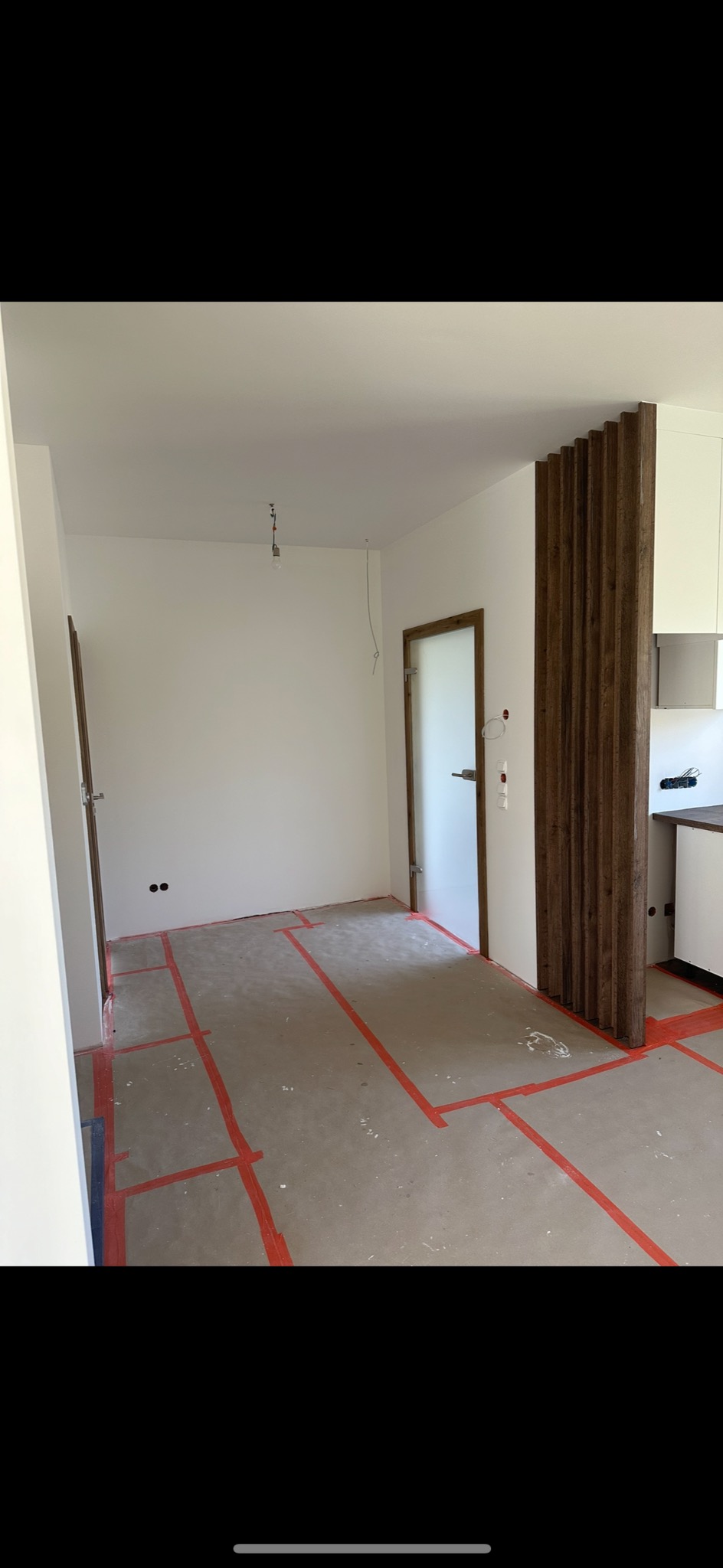 Wnętrze pokoju w trakcie remontu: ściany pomalowane na biało, podłoga z zaznaczonymi czerwonymi liniami, widoczne drzwi z drewnianą ramą i drewniana ścianka działowa.