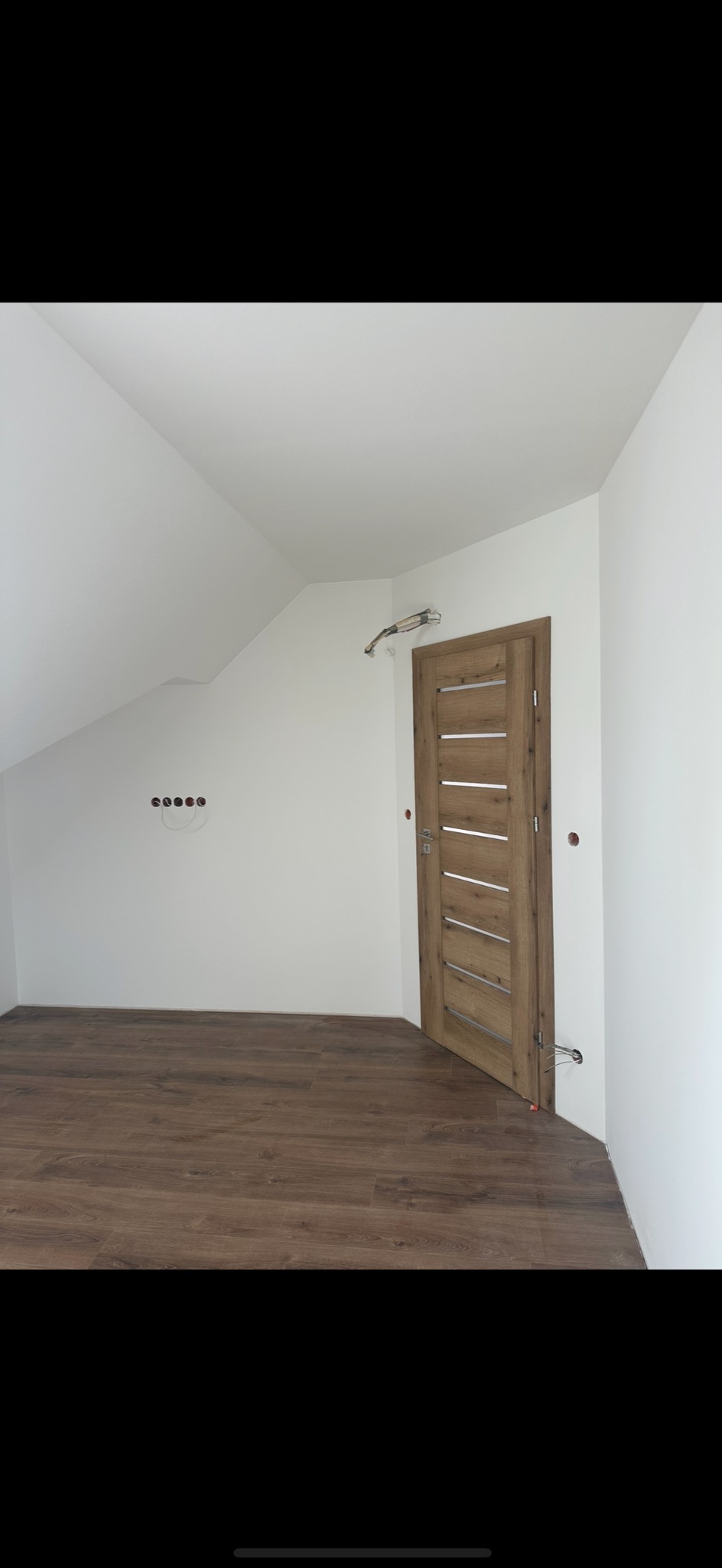 Poddasze po remoncie: świeżo pomalowane białe ściany, drewnopodobna podłoga, zamontowane gniazdka elektryczne i nowa drewniana, nowoczesna drzwi wewnętrzne z poziomymi przeszkleniami.