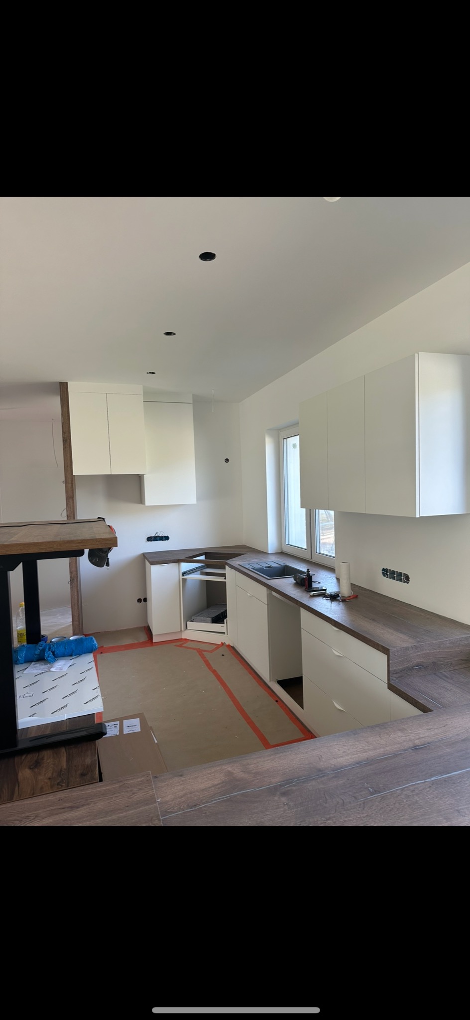 Wnętrze kuchni w trakcie montażu: białe szafki wiszące i dolne z blatem imitującym ciemne drewno, widoczne przygotowania do podłączenia instalacji elektrycznej i hydraulicznej, podłoga...