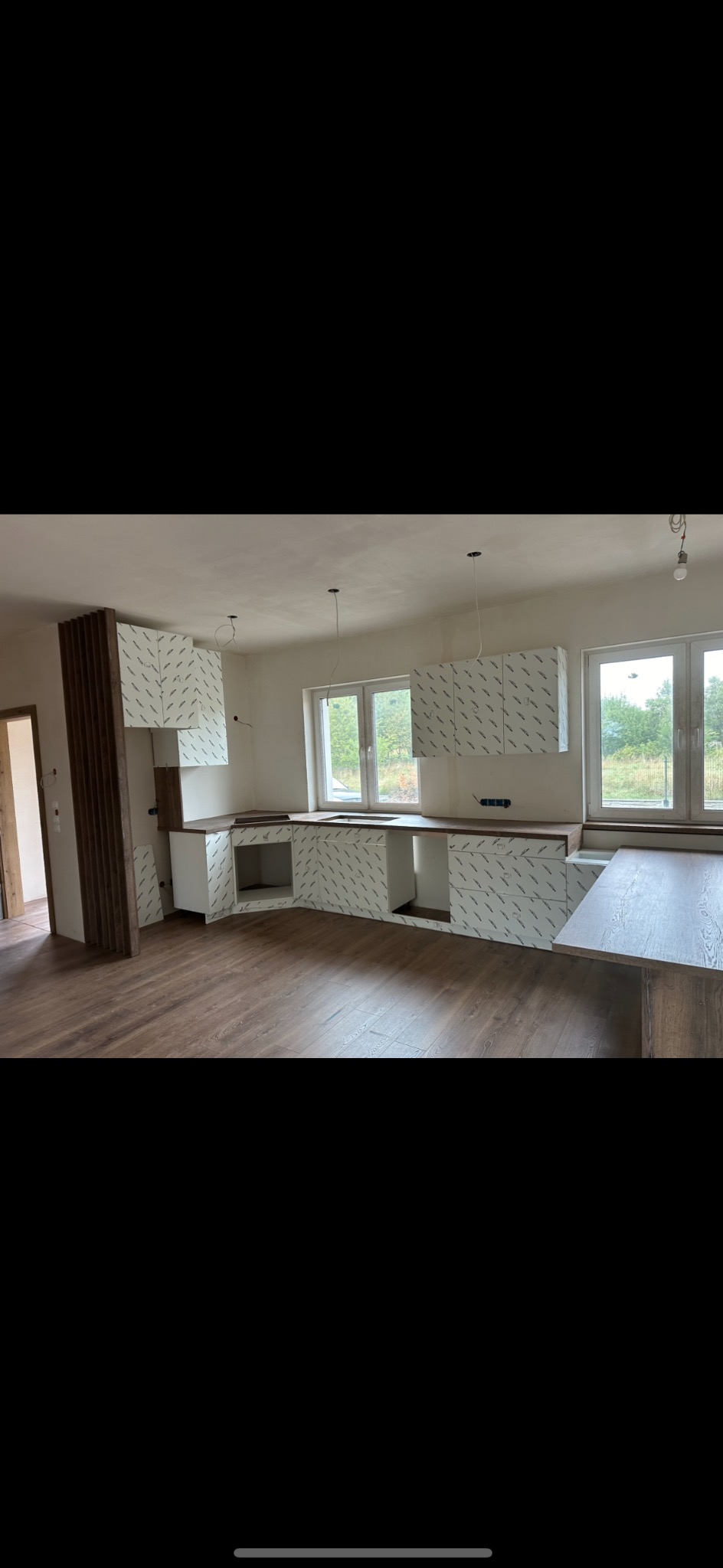 Wnętrze kuchni w trakcie montażu, widoczne szafki z folią ochronną, drewniany blat i podłoga, surowe ściany i wystające kable.