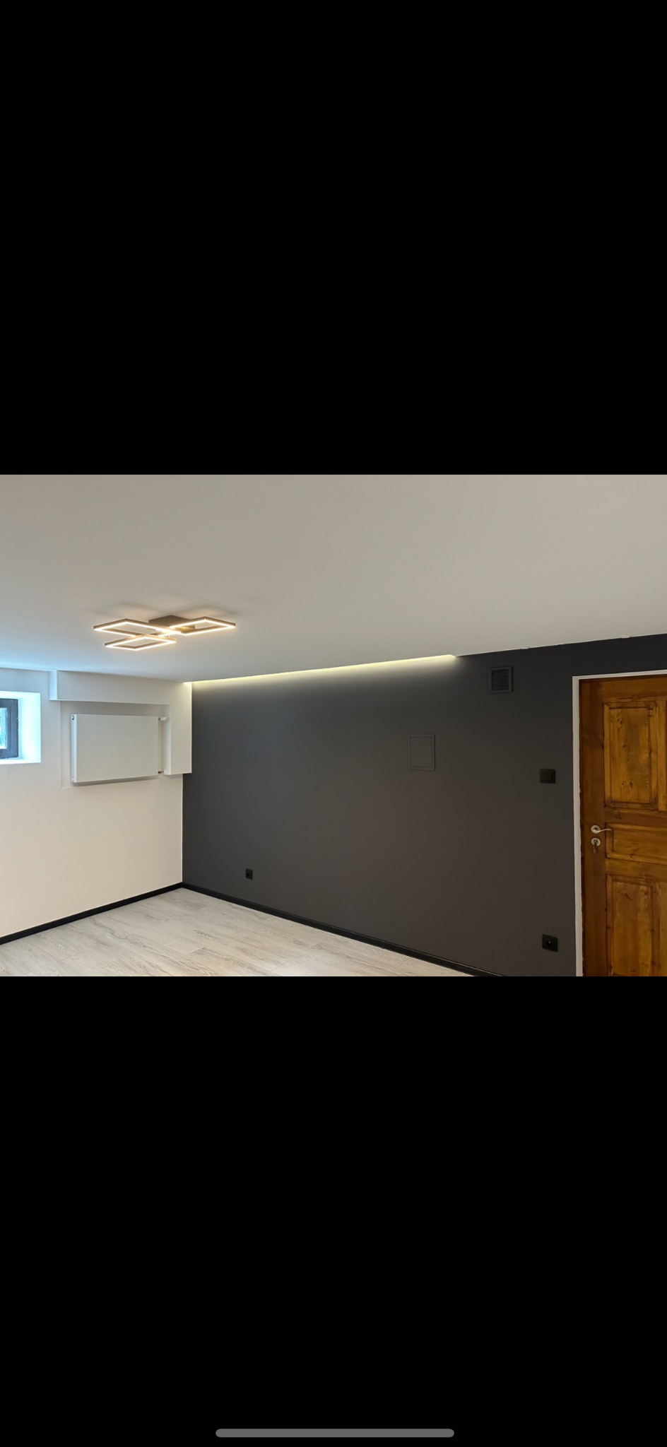 Wnętrze po remoncie: szara ściana z ukrytym oświetleniem LED pod sufitem, drewniane drzwi, jasna podłoga i nowoczesna lampa sufitowa.