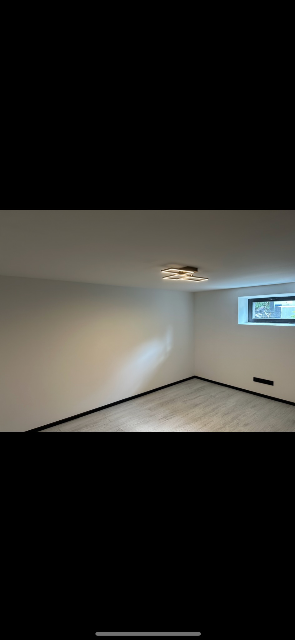 Pusty, świeżo wyremontowany pokój z białymi ścianami, panelami podłogowymi w odcieniach szarości i nowoczesną, kwadratową lampą sufitową.