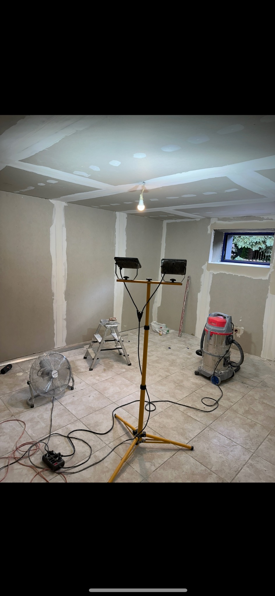 Pokój w trakcie prac wykończeniowych: ściany z płyt gipsowo-kartonowych ze szpachlowanymi łączeniami, halogen na statywie, wentylator, odkurzacz przemysłowy i drabina.