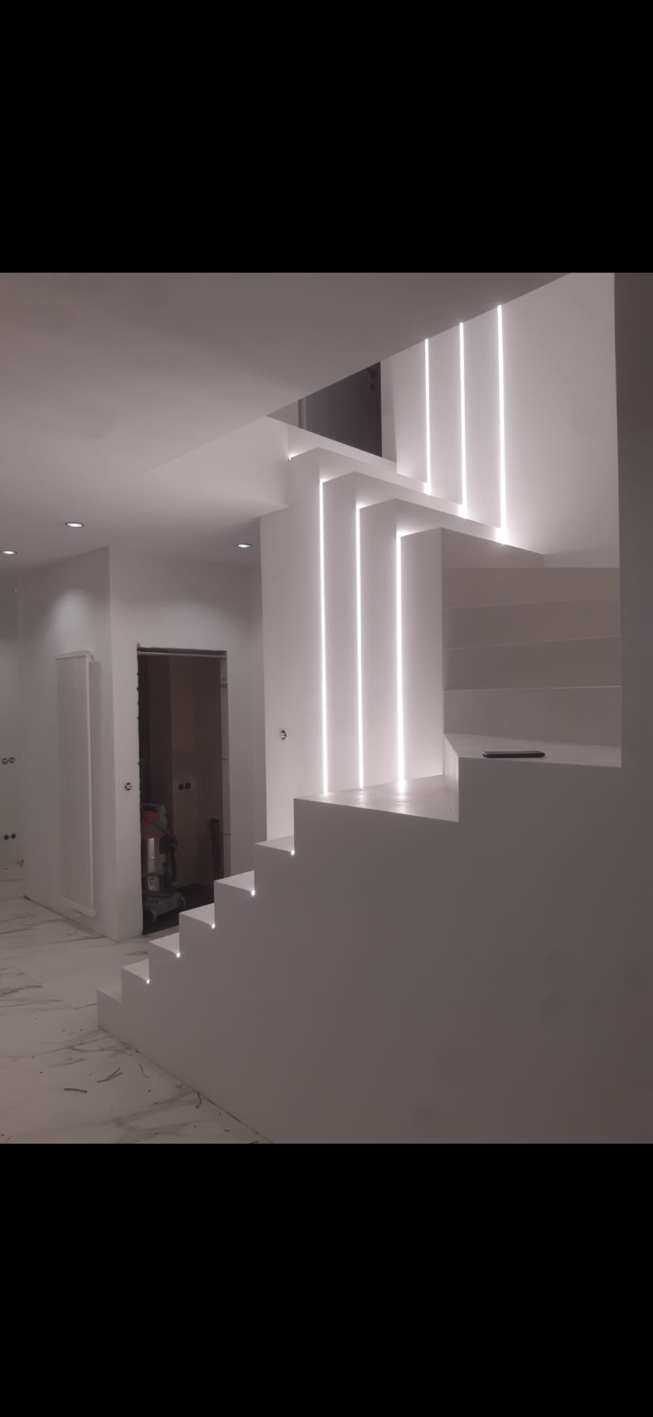 Nowoczesne, białe schody z podświetleniem LED w pionowych listwach, widok na wykończone wnętrze z grzejnikiem i otwartym przejściem do innego pomieszczenia.