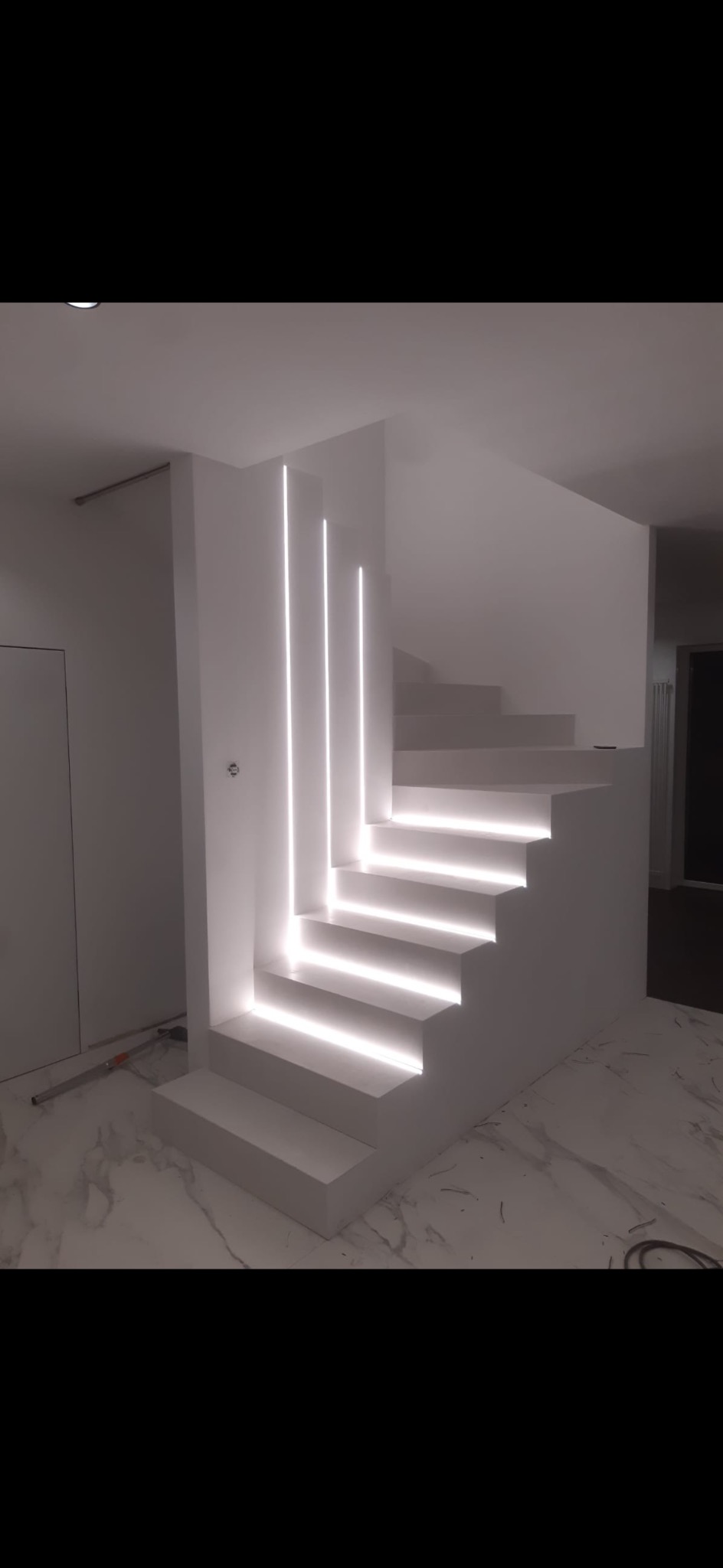 Minimalistyczne, białe schody z podświetleniem LED wzdłuż stopni i pionowych paneli, marmurowa podłoga, widoczne narzędzia malarskie przy ścianie.