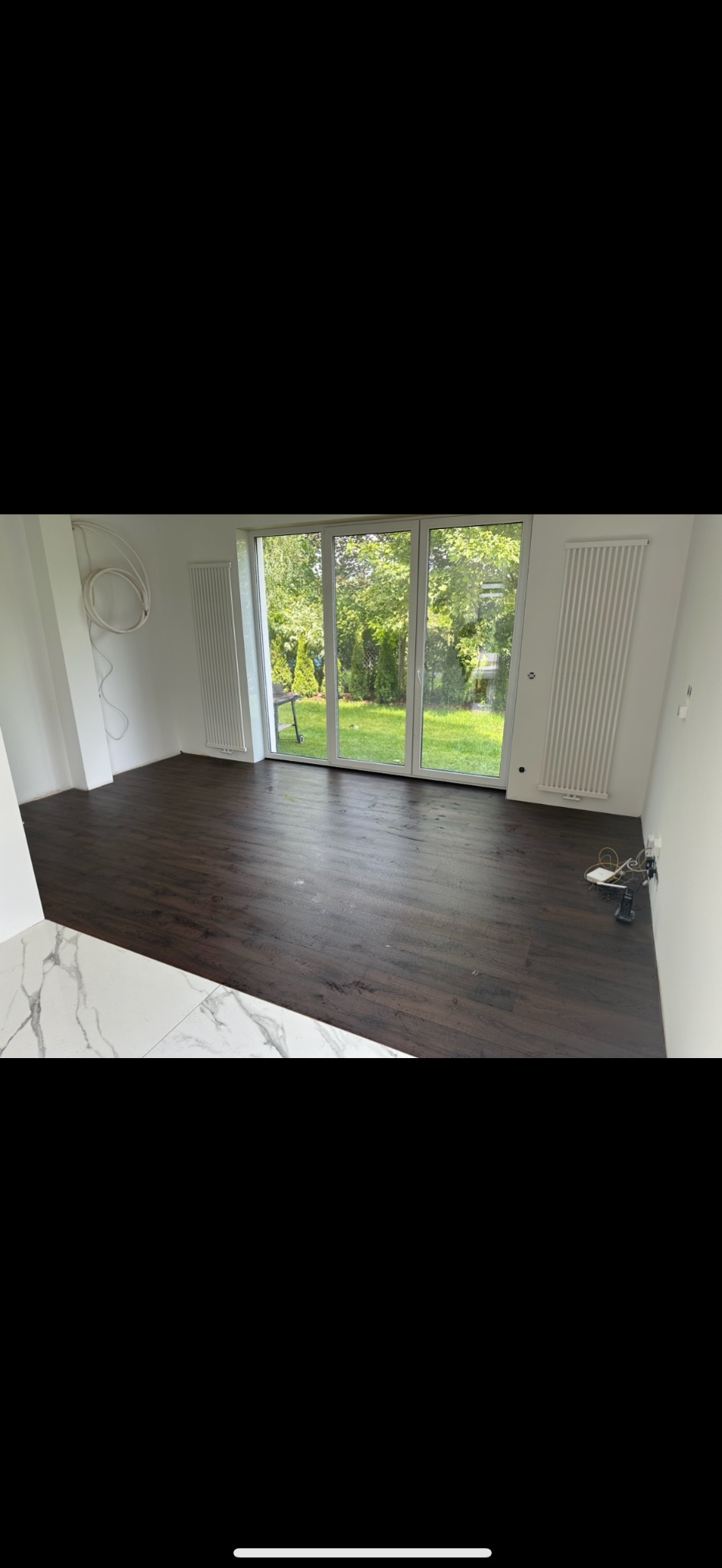 Nowo wykończone wnętrze z ciemną podłogą imitującą drewno, białymi ścianami, grzejnikami panelowymi i dużymi przesuwnymi drzwiami balkonowymi z widokiem na zielony ogród.