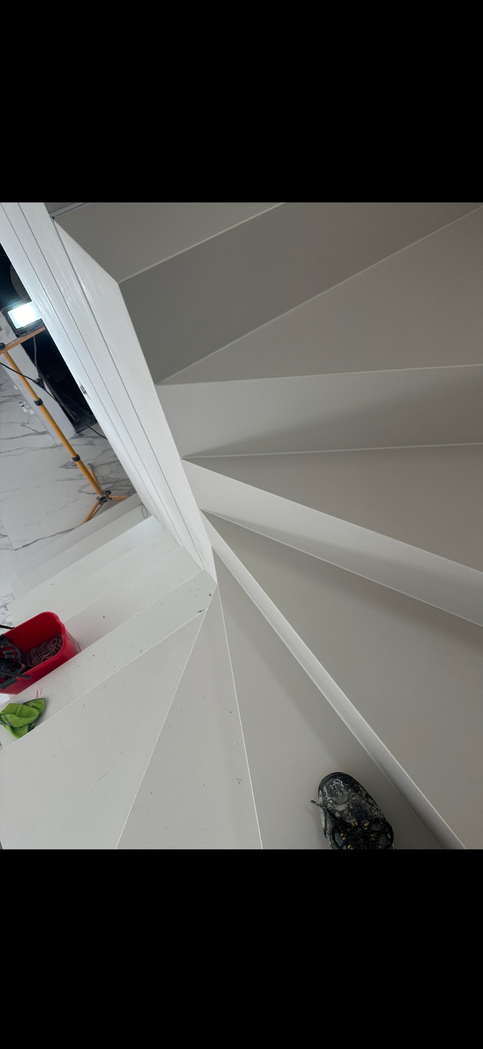 Wysoki kąt widzenia na spiralne, pomalowane na biało schody z widocznym sprzętem oświetleniowym, czerwonym wiadrem i butem roboczym.