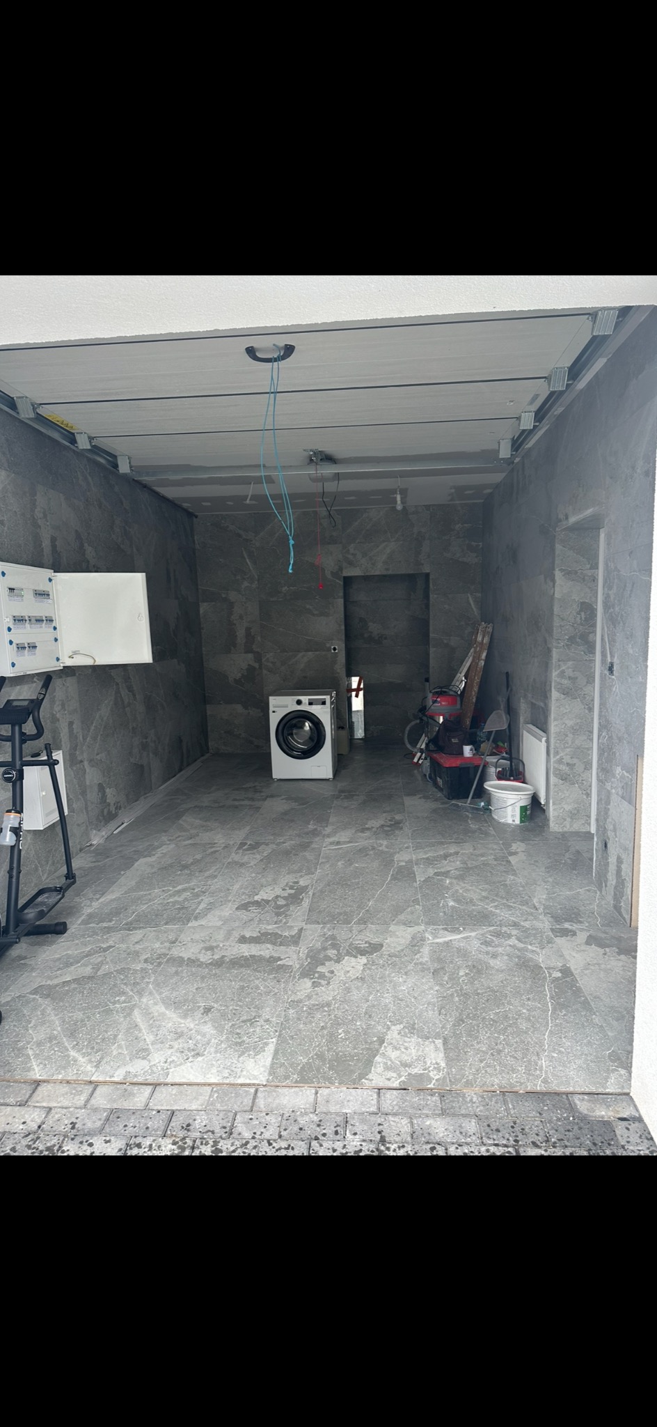 Wnętrze pomieszczenia w trakcie remontu: szare płytki na podłodze i ścianach, częściowo zamontowany sufit podwieszany, pralka, drabina i narzędzia w tle.