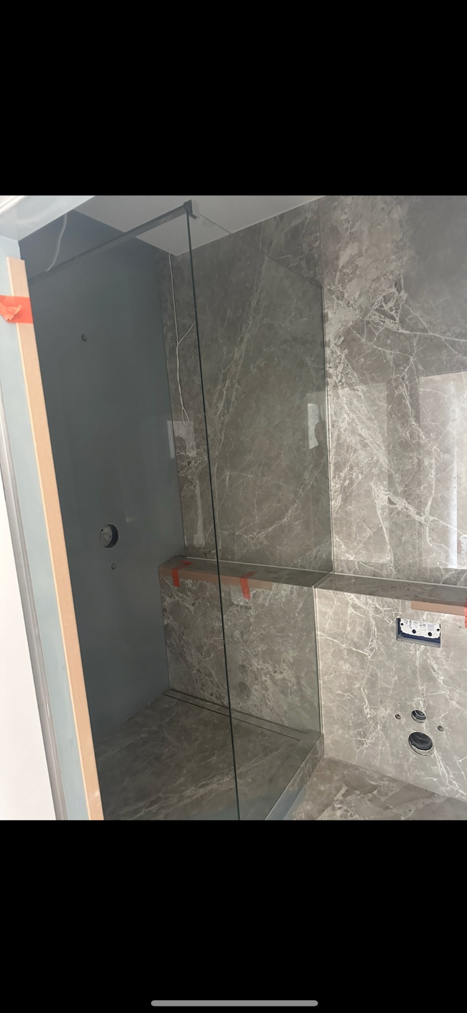 Szklana kabina prysznicowa z szarymi marmurowymi płytkami, widoczne otwory na armaturę, zabezpieczona taśmą malarską na czas prac wykończeniowych.