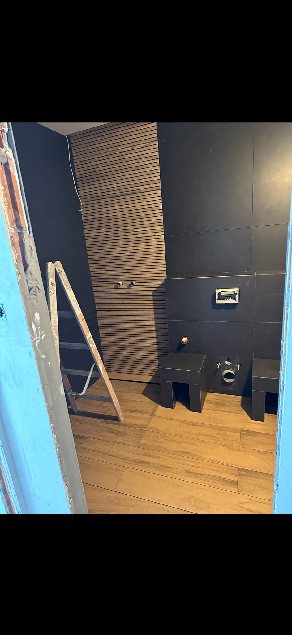 Łazienka w trakcie wykańczania: drewnopodobna podłoga, czarne płytki na ścianach, drewniane panele ścienne, drabina oparta o ścianę, widok zza ościeżnicy.