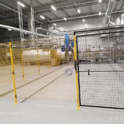 Widok hali przemysłowej z instalacją rur, zbiorników i metalowych siatek ochronnych z żółtymi słupkami, z widocznymi elementami wentylacji pod sufitem.