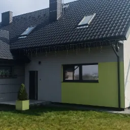 Widok z zewnątrz na ocieplony dom z ciemną dachówką, oknami i elementami elewacji w odcieniach szarości, bieli i limonki. Na elewacji widoczne ozdobne światełka.