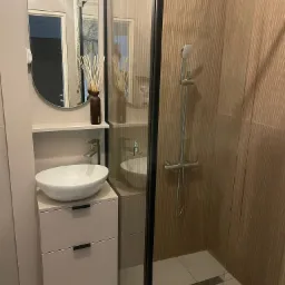 Nowoczesna łazienka z kabiną prysznicową z czarnymi ramami, umywalką nablatową na szafce z szufladami i dekoracyjnym dyfuzorem zapachowym na półce.