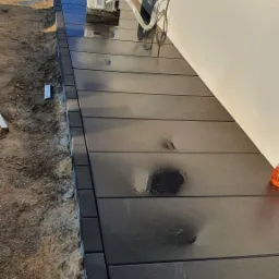 Świeżo ułożona, mokra, grafitowa kostka brukowa przy ścianie budynku, z widoczną instalacją klimatyzacji i niezakończonym obrzeżem z piaskiem.