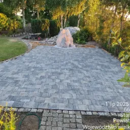 Ułożona szara kostka brukowa na posesji, widoczne przygotowania do dalszych prac, w tle zieleń drzew i krzew&oacute;w, mała architektura ogrodowa.