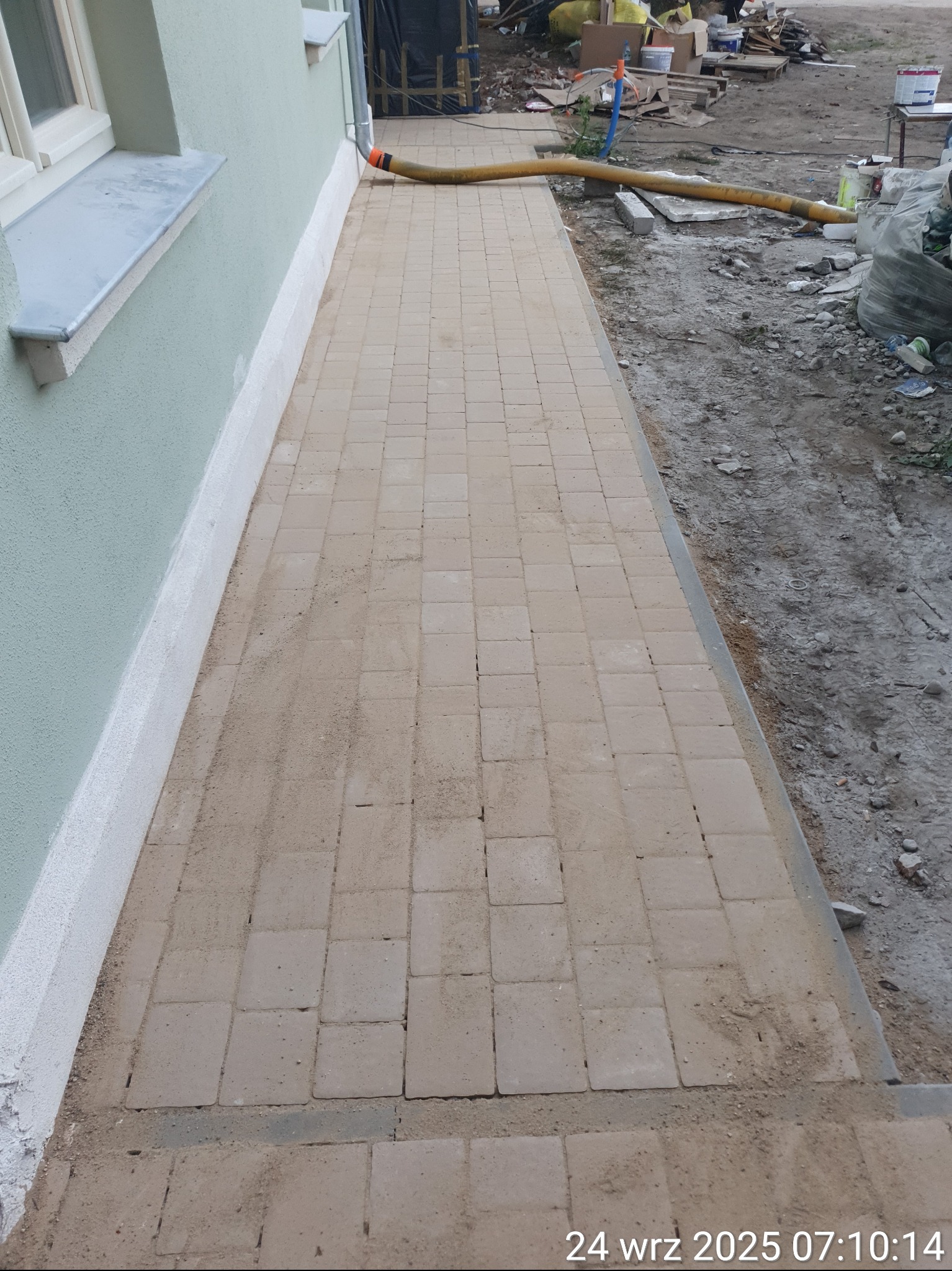 Ścieżka z beżowej kostki brukowej przy budynku, z widocznymi śladami piasku i prac wykończeniowych. W tle widoczne materiały budowlane i narzędzia.