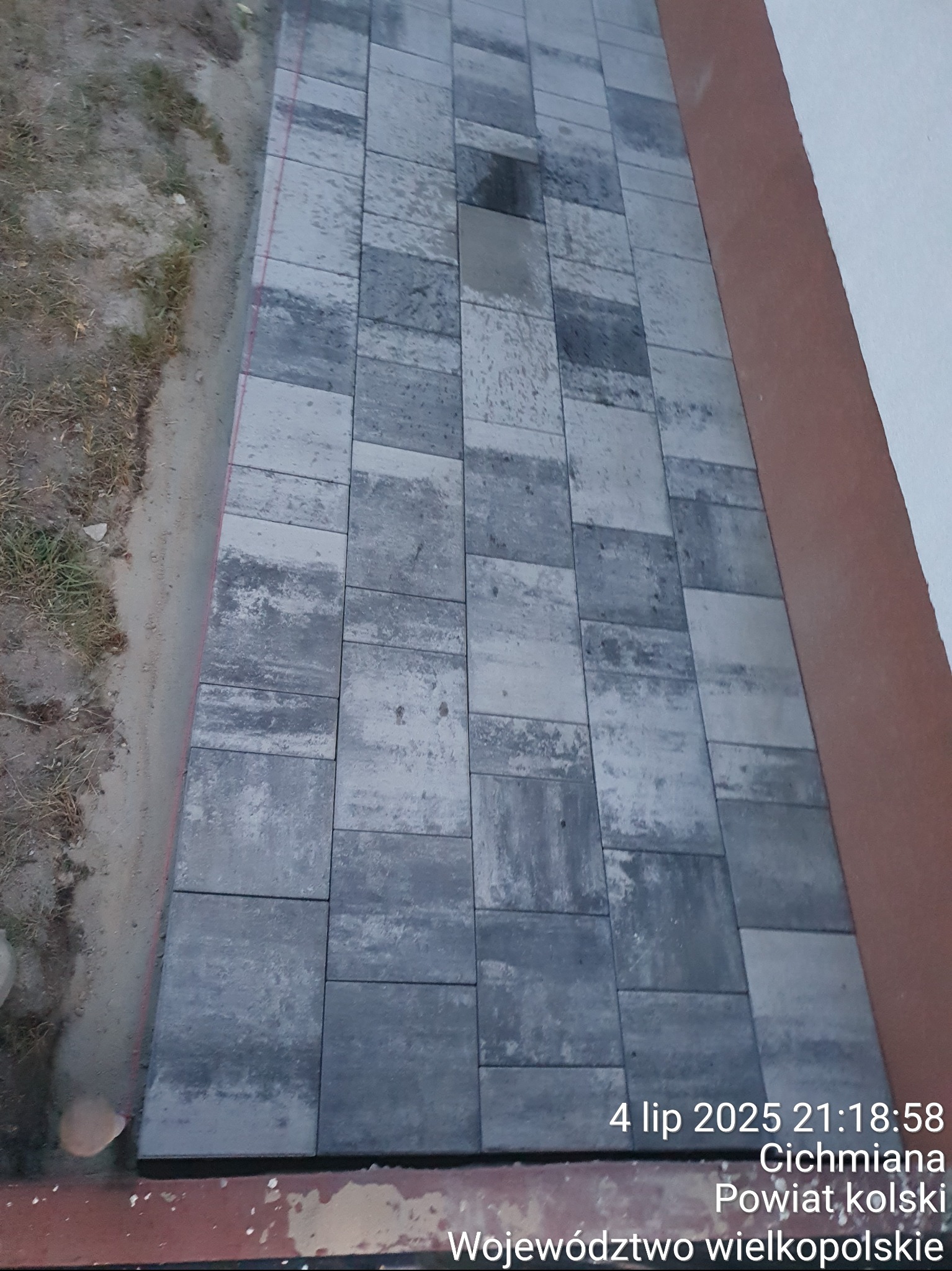 Ułożona, szara kostka brukowa tworząca ścieżkę przy budynku. Widoczna linia oddzielająca kostkę od ziemi. Zdjęcie z datą i lokalizacją w rogu.