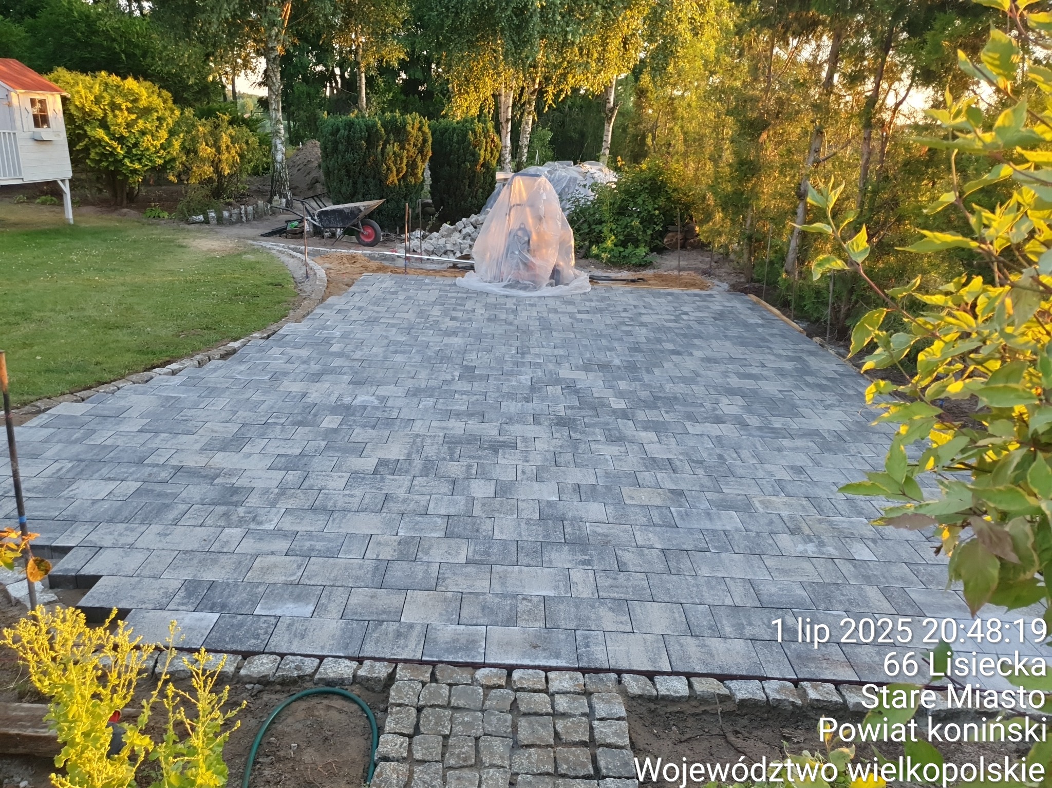 Ułożona szara kostka brukowa na posesji, widoczne przygotowania do dalszych prac, w tle zieleń drzew i krzewów, mała architektura ogrodowa.