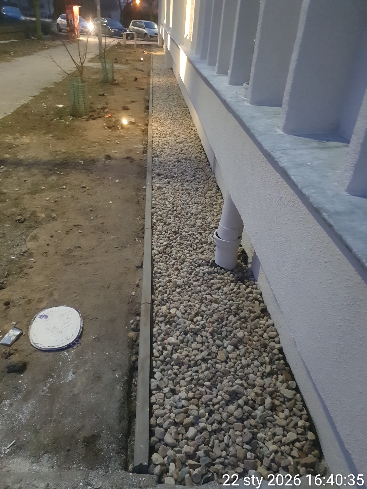 Wykończona rabata z kamykami przy budynku, oddzielona obrzeżem. Widoczny fragment elewacji i chodnika. Prace brukarskie w przestrzeni miejskiej.