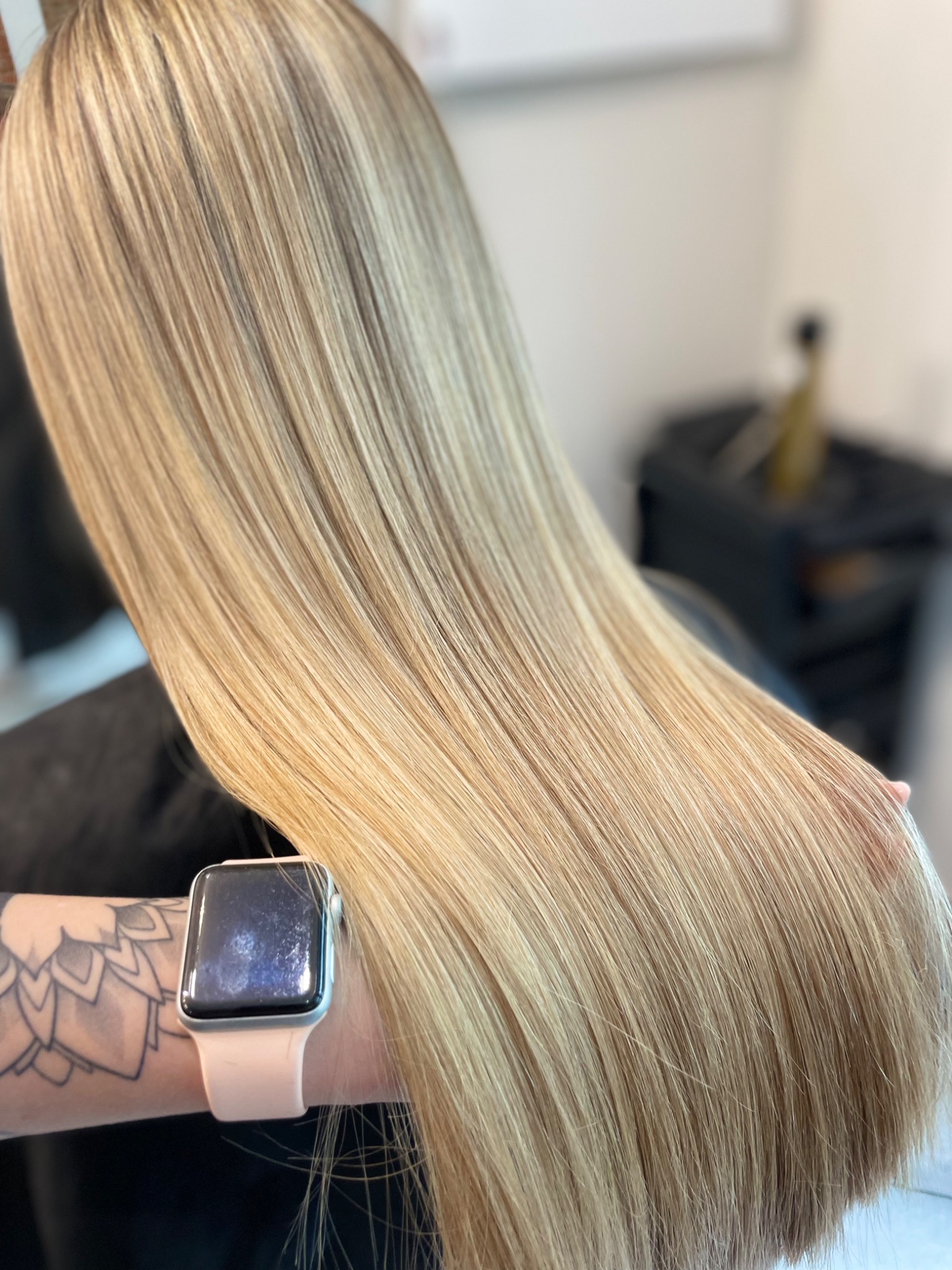Długie, proste, blond włosy z delikatnymi refleksami, trzymane przez wytatuowaną rękę z zegarkiem Apple Watch.