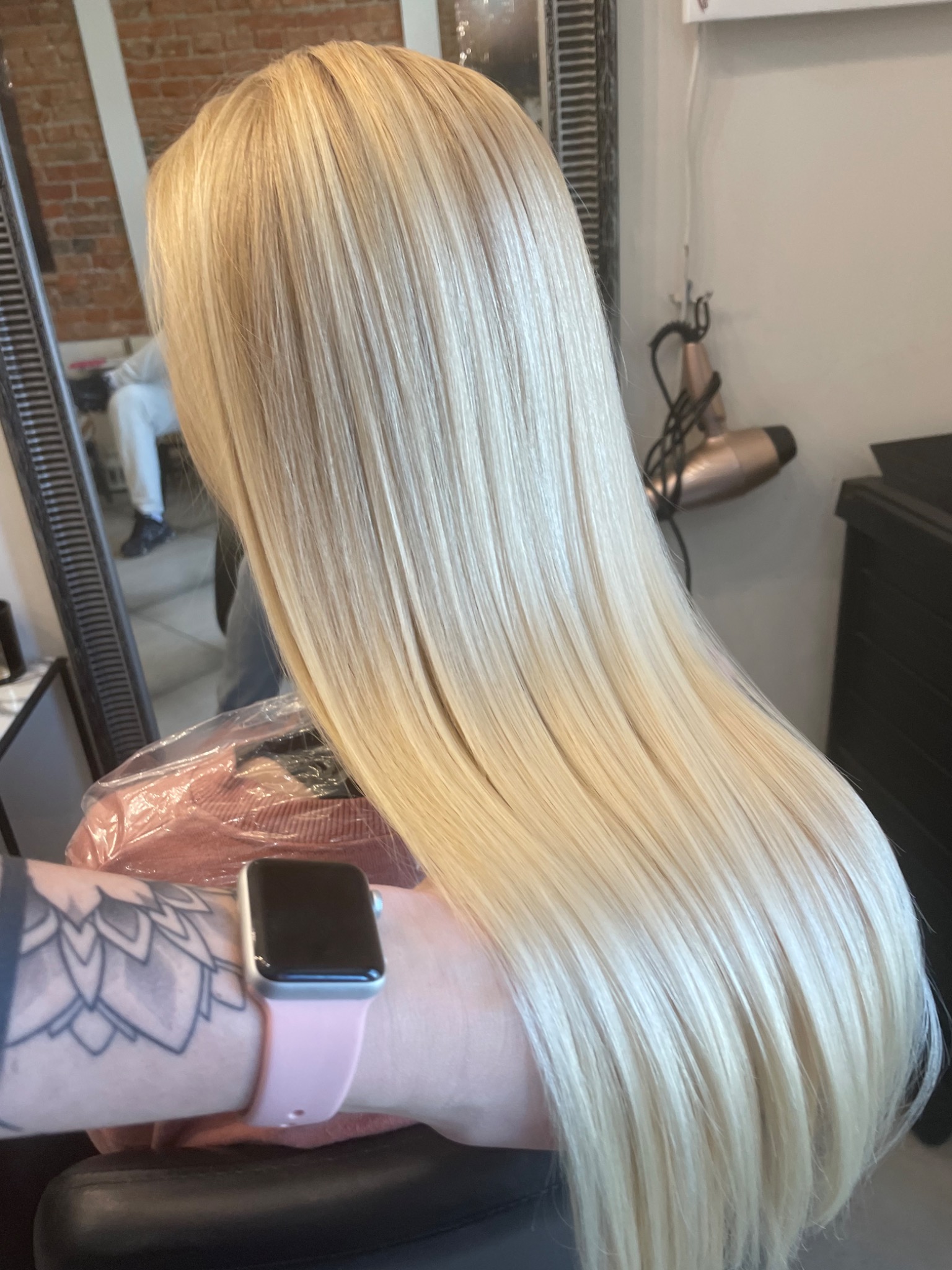 Długie, proste, platynowe blond włosy po koloryzacji, widoczne z tyłu głowy, z ręką i zegarkiem na pierwszym planie.