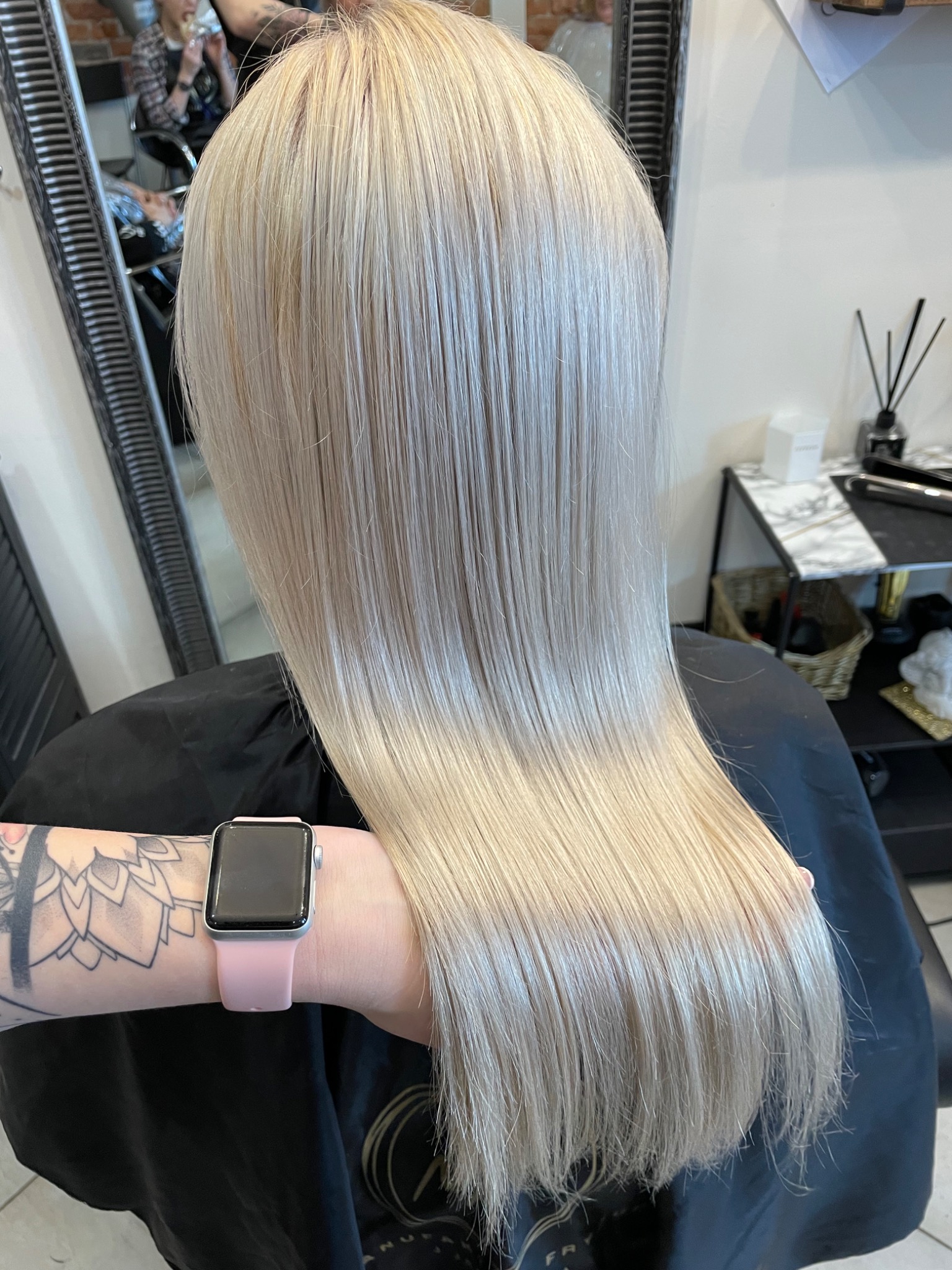 Świeżo po koloryzacji platynowy blond, długie proste włosy, widoczny fragment ramienia z tatuażem i smartwatchem.