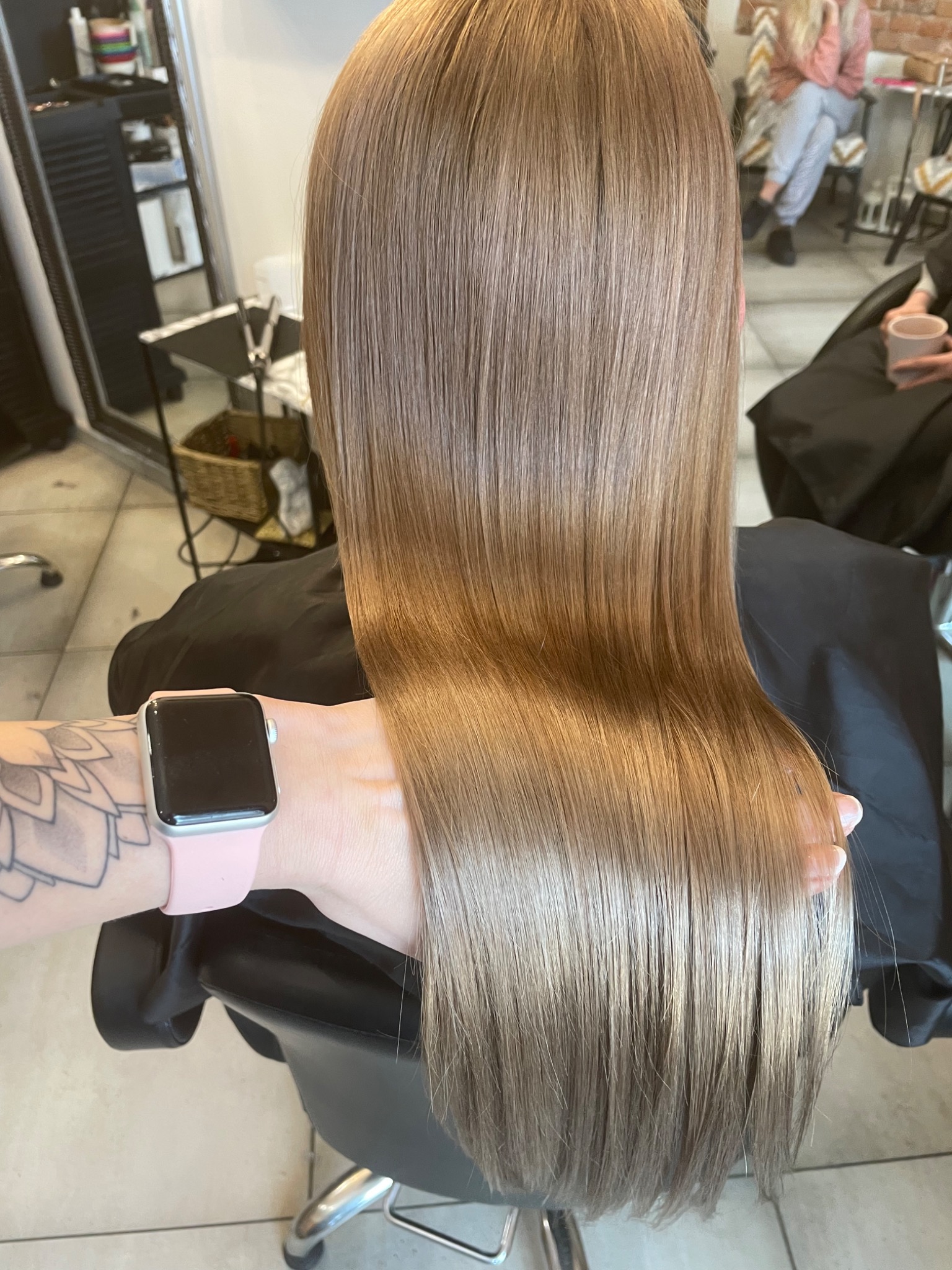 Proste, lśniące włosy w odcieniach blondu i brązu, widok z tyłu, ręka z tatuażem i smartwatchem przytrzymuje pasmo włosów.