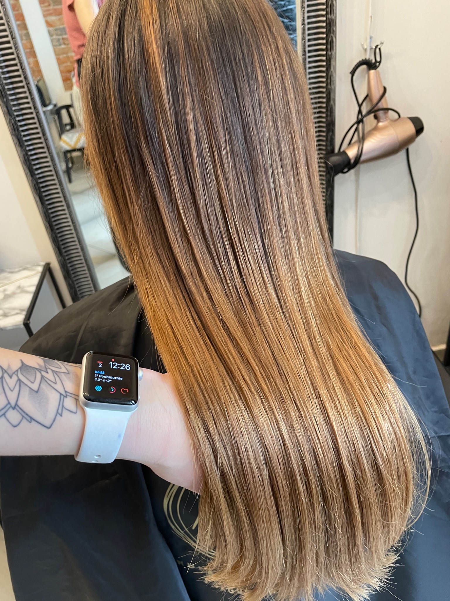 Długie, proste włosy z refleksami w odcieniach blondu i brązu, widoczne z tyłu głowy, na ramieniu widoczny tatuaż i smartwatch.