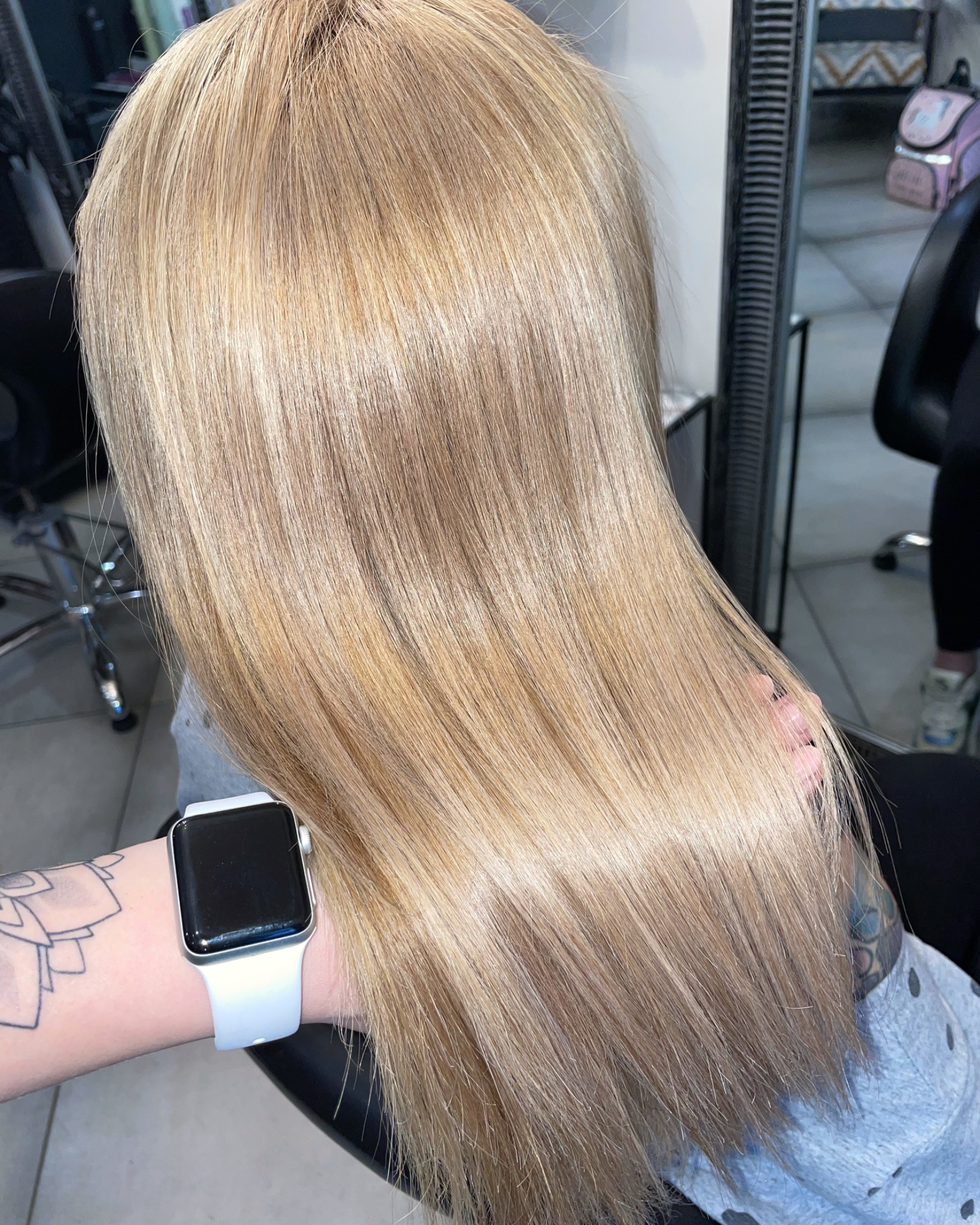 Świeżo ufarbowane, lśniące, proste włosy w odcieniu jasnego blondu, widok z tyłu, na nadgarstku widoczny smartwatch z białym paskiem i tatuaż.