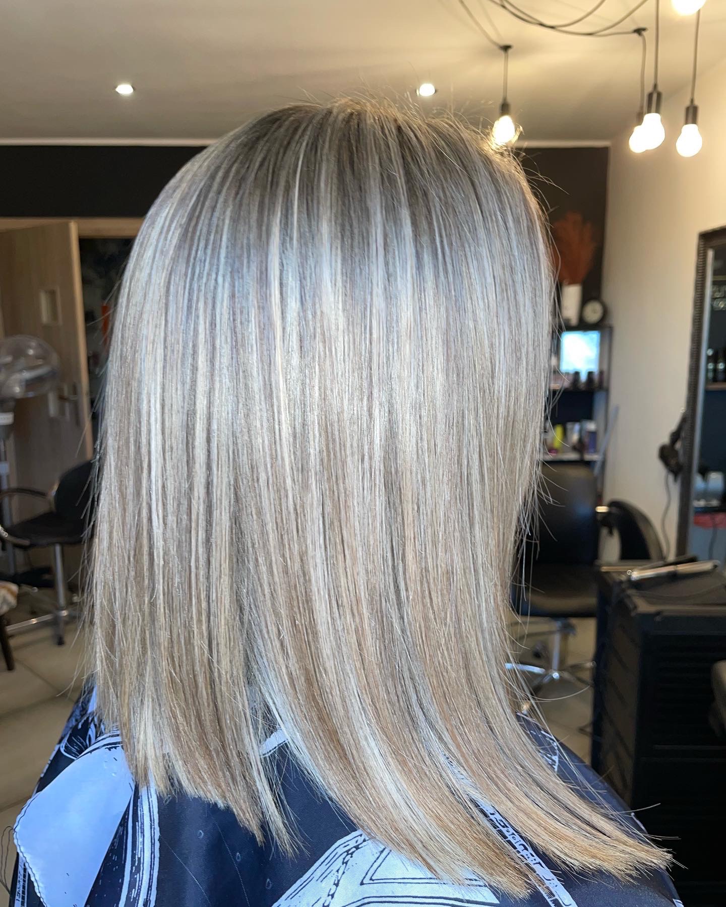 Włosy blond z pasemkami, ścięte prosto do ramion, widok z tyłu, klientka w pelerynie ochronnej w salonie.
