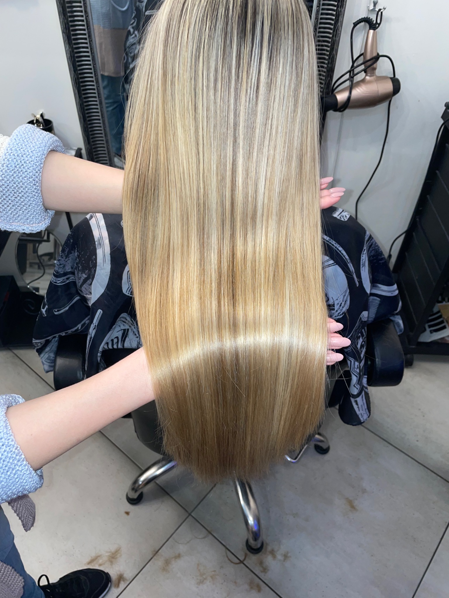 Długie, blond włosy z ciemniejszymi odrostami, po zabiegu prostowania, trzymane przez dłonie z delikatnym manicure, widoczny fragment fryzjerskiego fotela i podłogi z obciętymi włosami.