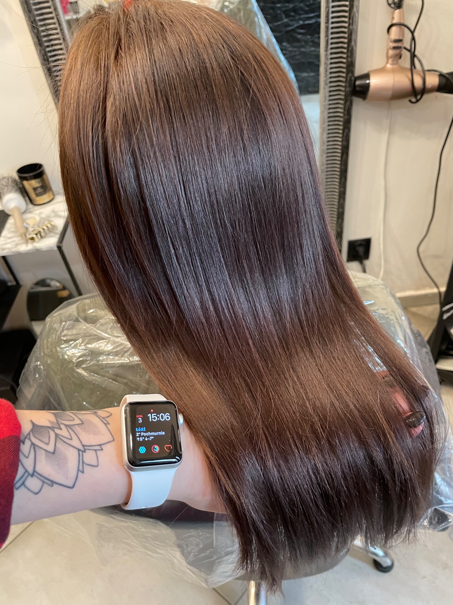 Świeżo ufarbowane, lśniące brązowe włosy, widoczne od tyłu, z delikatnym ombre na końcach, w tle suszarka do włosów i akcesoria na półce, na pierwszym planie ręka z tatuażem i smartwatchem.