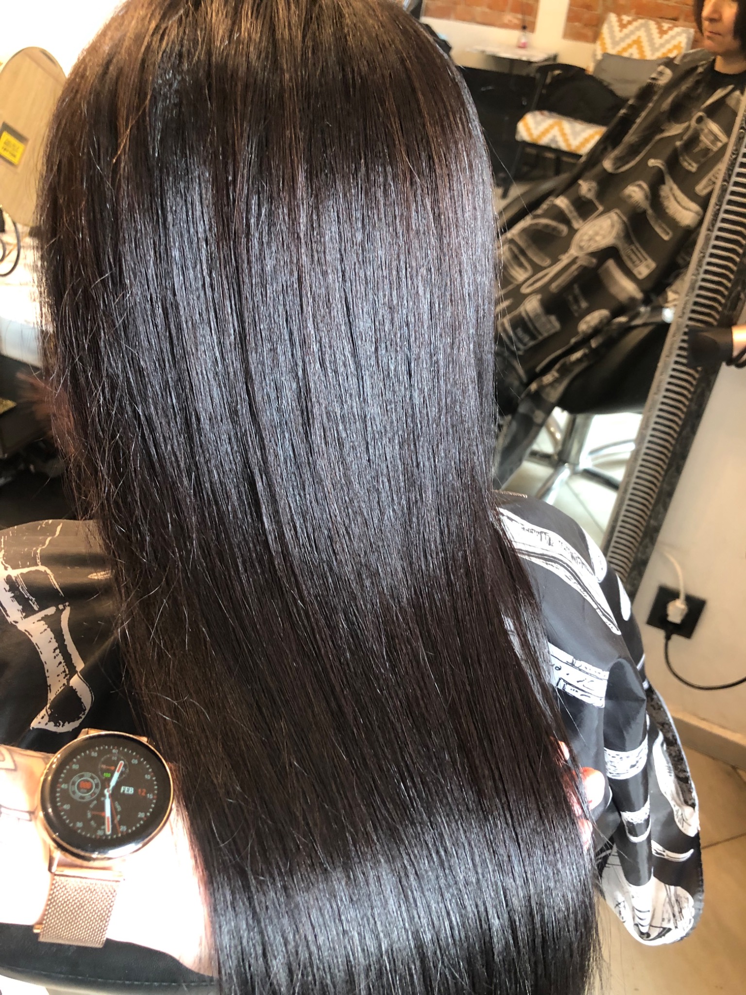 Ciemne, lśniące, wyprostowane włosy po zabiegu fryzjerskim, widoczne z tyłu, klientka w pelerynie fryzjerskiej z zegarkiem na ręce.