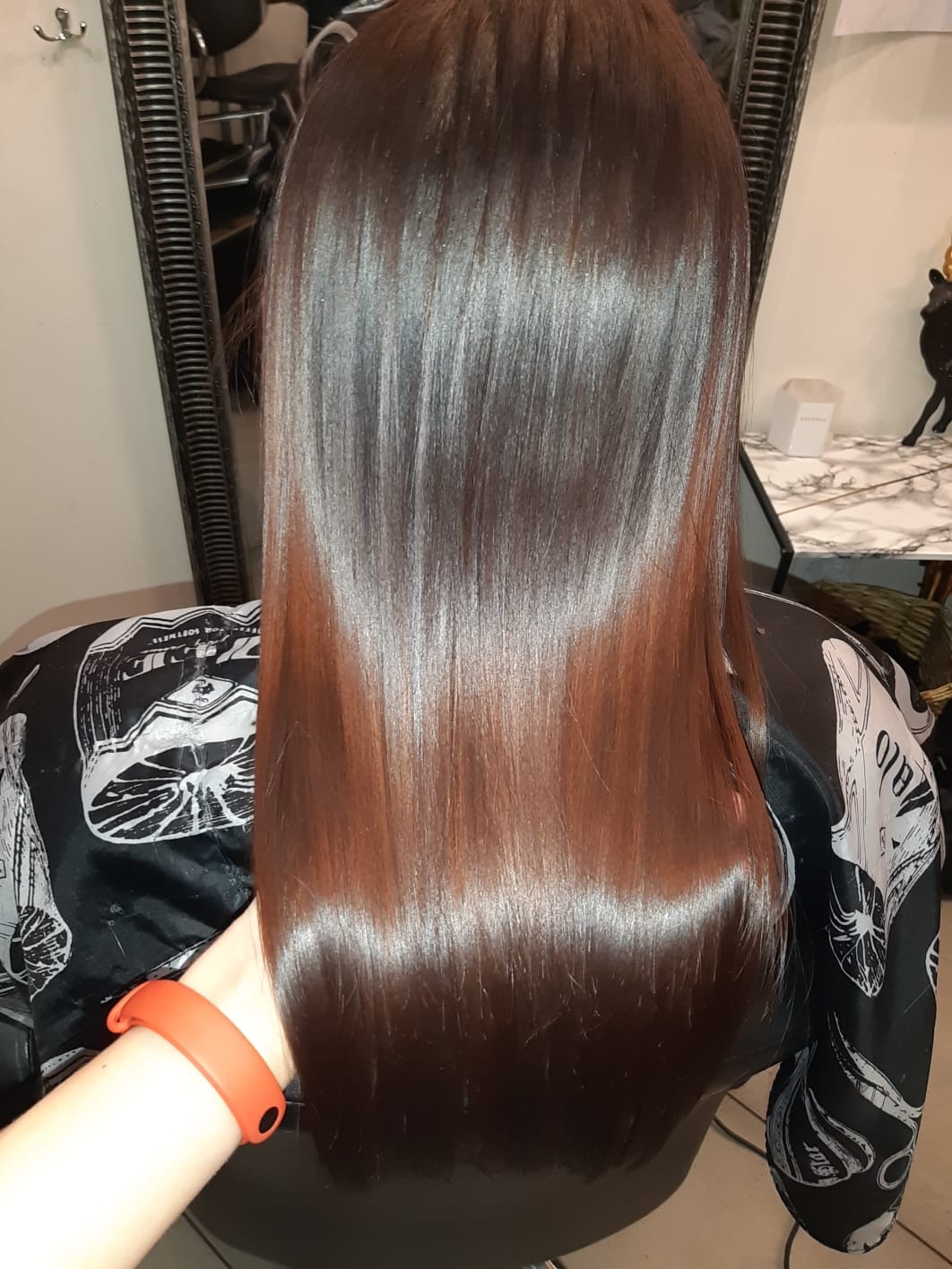 Wypielęgnowane, długie, brązowe włosy z refleksami, widoczne od tyłu, nałożony czarny fartuch ochronny z białym wzorem, pomarańczowa bransoletka na nadgarstku