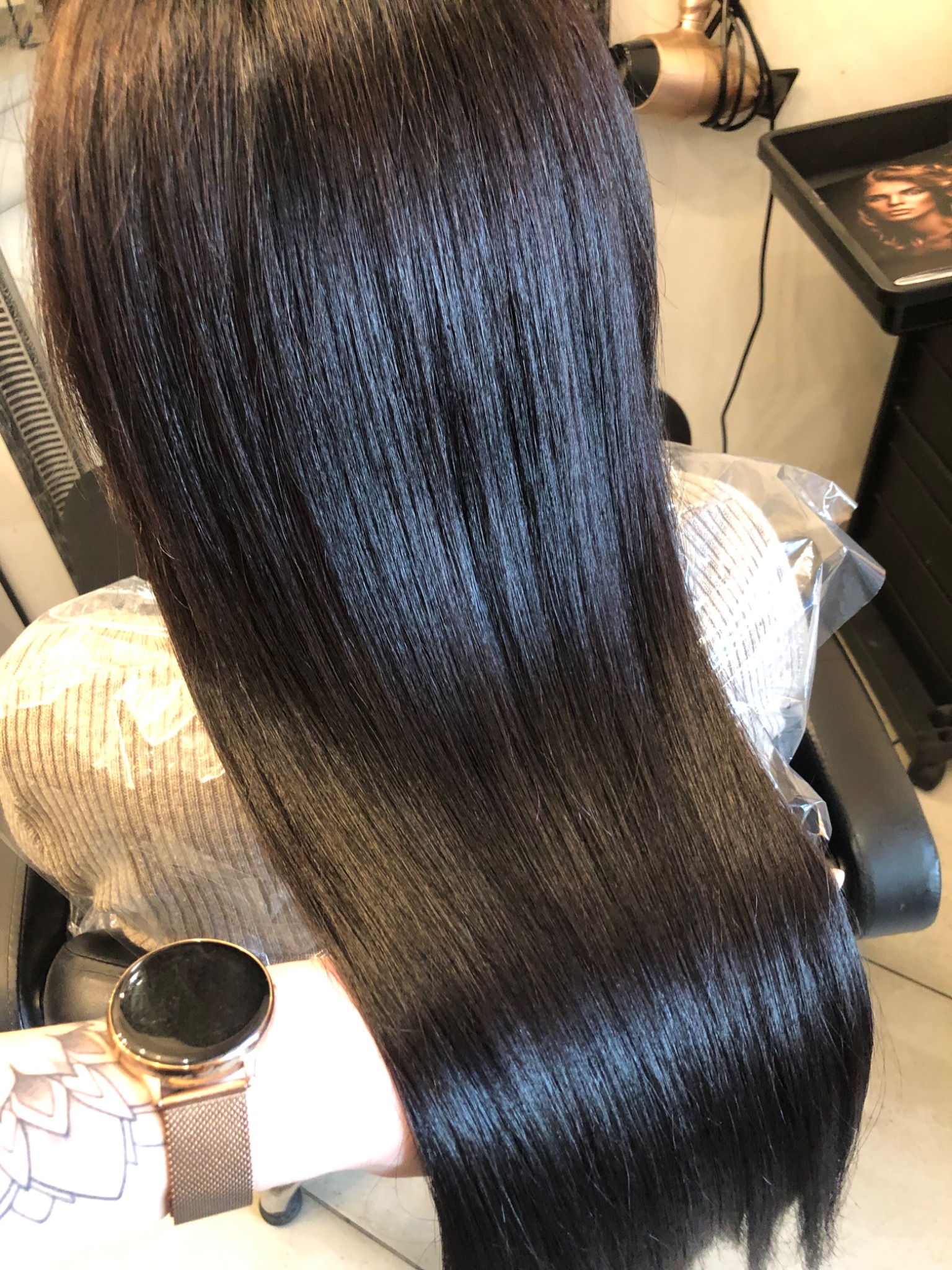 Błyszczące, ciemne, proste włosy po zabiegu prostowania keratynowego, widoczne z tyłu głowy, z bliska, z zegarkiem na nadgarstku w kadrze.