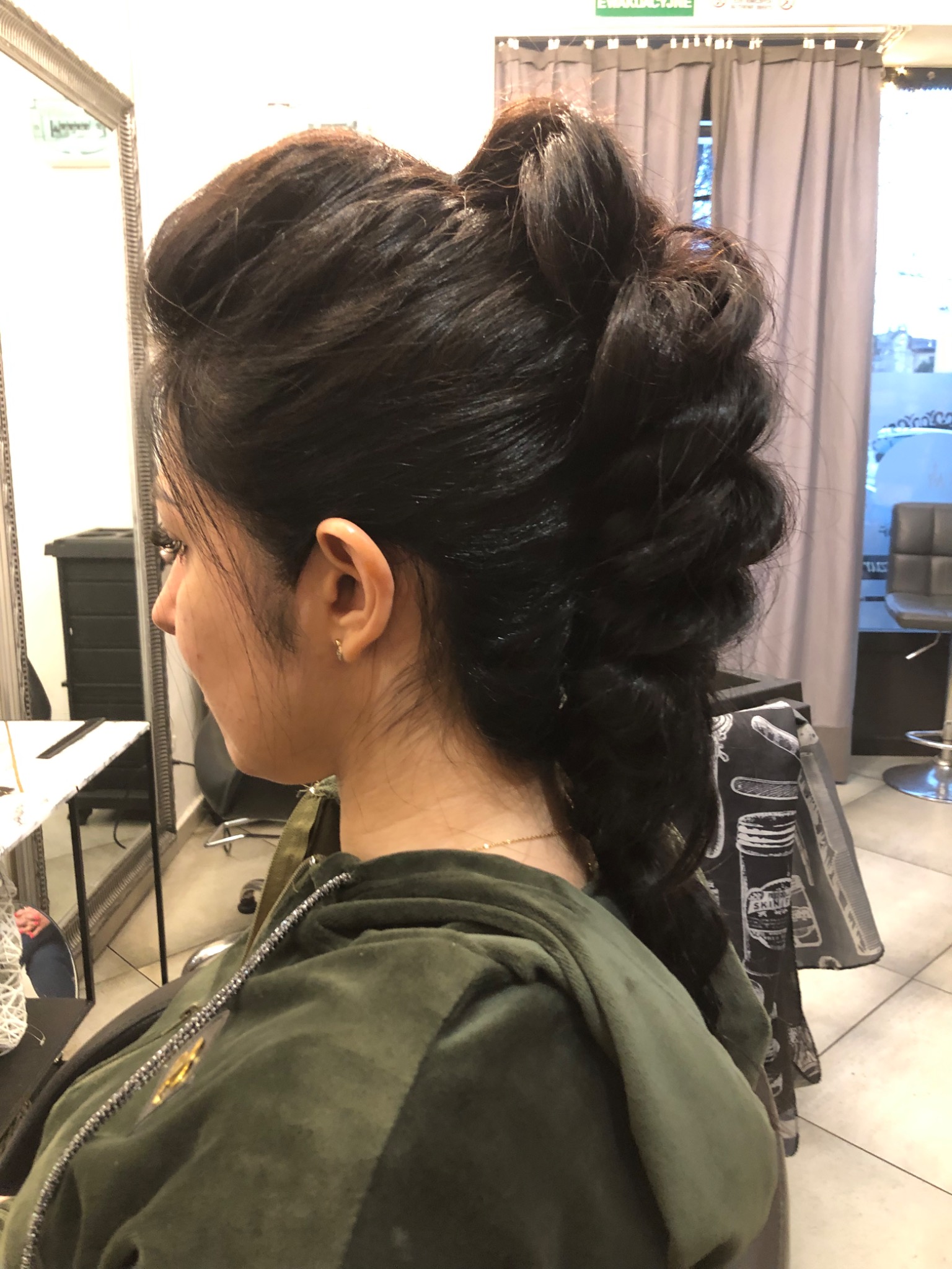 Elegancka fryzura upięta z ciemnych włosów, widok z boku na tle salonu z lustrem i zasłonami.