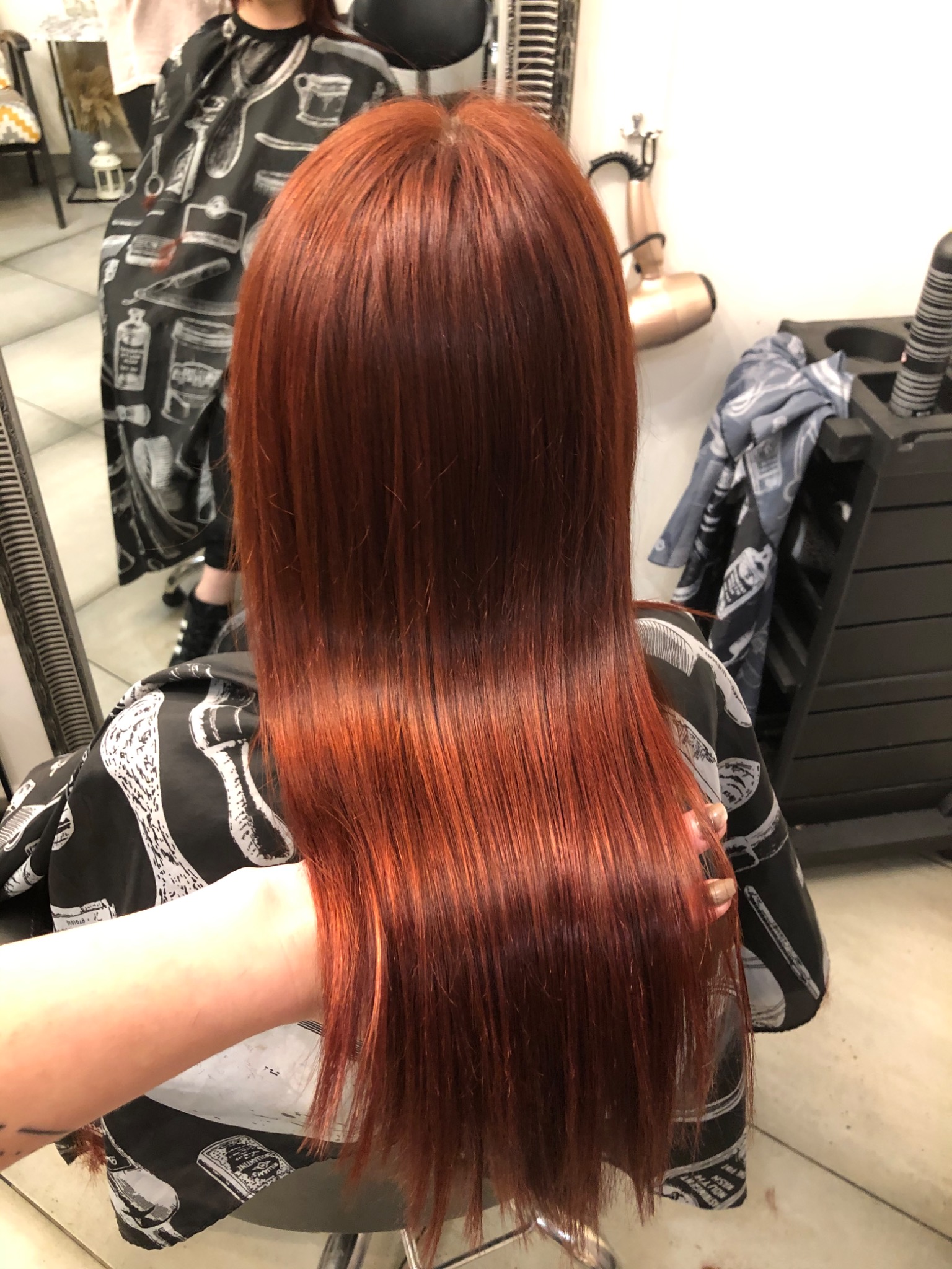 Proste, lśniące włosy w odcieniu miedzianej czerwieni, widok z tyłu, klientka w pelerynie ochronnej z motywem fryzjerskim.
