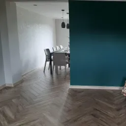 Salon z podłogą ułożoną w jodełkę, ściana pomalowana na butelkową zieleń, w tle widoczna jadalnia z szarą tapetą w delikatny wzór.