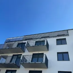 Nowoczesny budynek mieszkalny z balkonami zabezpieczonymi czarnymi, metalowymi balustradami na tle błękitnego nieba.