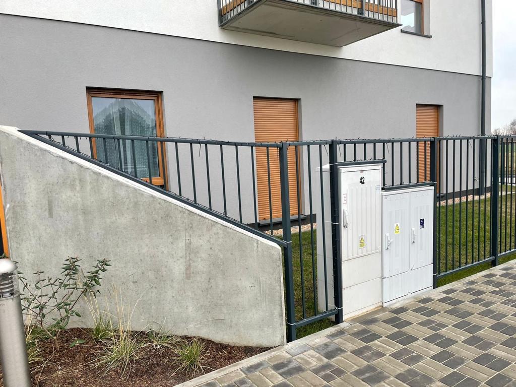 Nowoczesne ogrodzenie z pionowych prętów w kolorze grafitowym, zabezpieczające teren przy budynku z szarą elewacją i brązowymi roletami okiennymi, z widocznymi skrzynkami elektrycznymi obok bramy.
