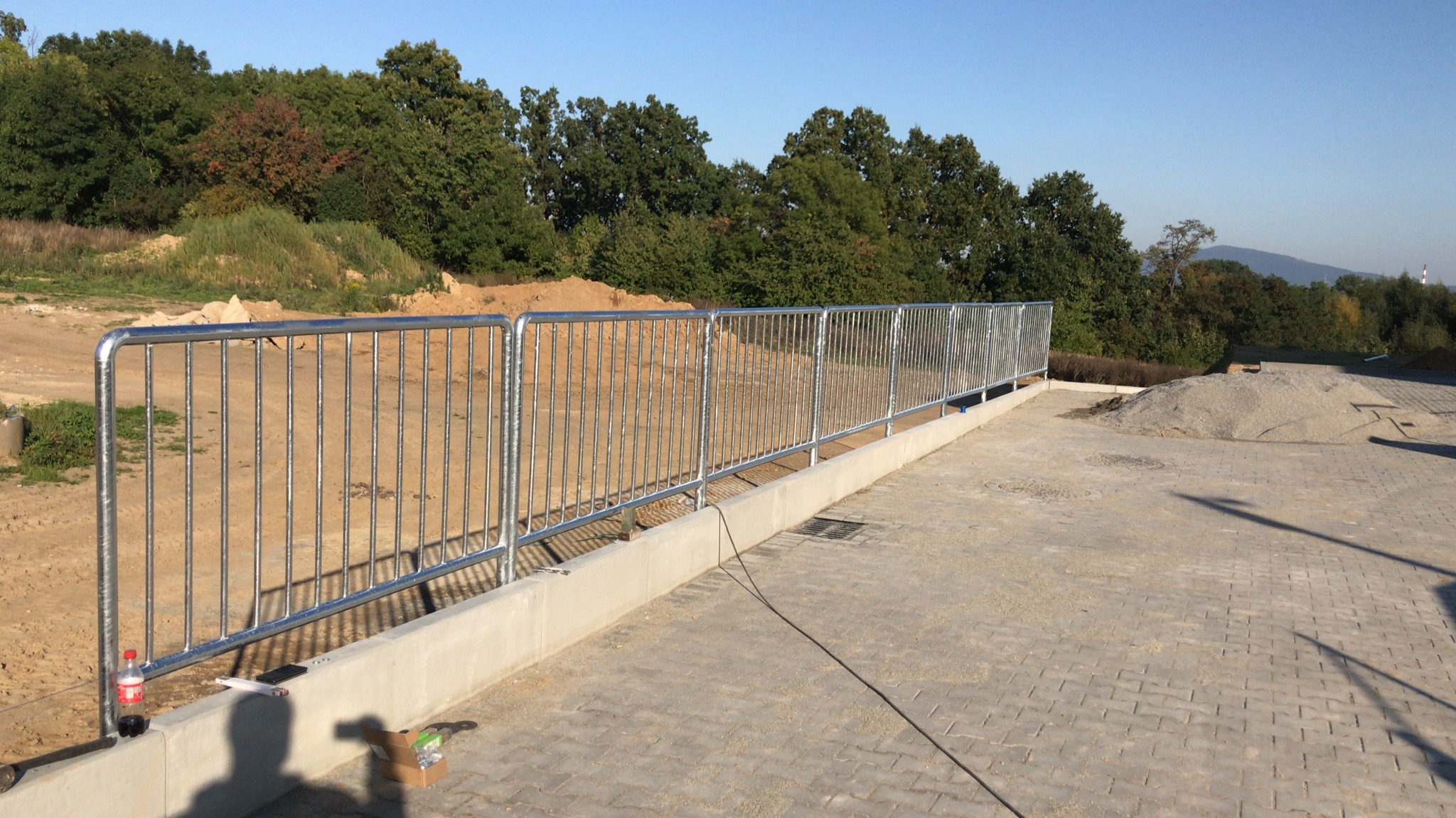 Nowo zamontowane, ocynkowane zabezpieczenie barierowe na betonowej podstawie, z widocznym kablem zasilającym, butelką napoju i narzędziami w tle placu budowy.