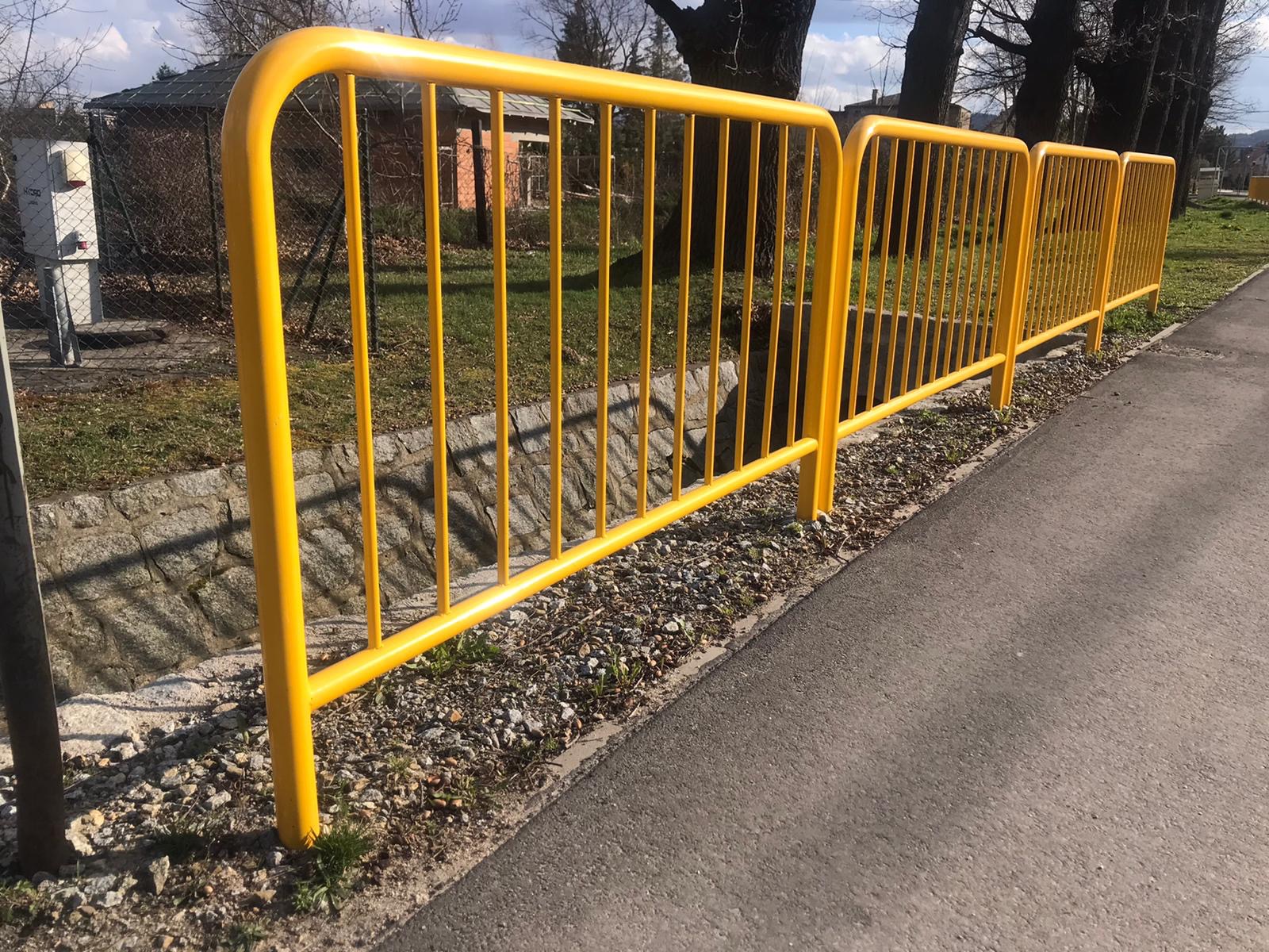 Żółte, metalowe barierki ochronne oddzielające chodnik od terenu zielonego, widoczne tło z budynkami i drzewami.