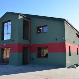 Nowoczesny budynek przemysłowy w kolorze zieleni i czerwieni z dużymi oknami i utwardzonym placem przed wejściem.