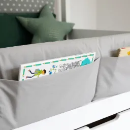 Szary, materiałowy organizer z kieszeniami na książki zawieszony na białym łóżku dziecięcym, widoczne książki z kolorowymi ilustracjami.