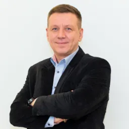 Portret mężczyzny w czarnej marynarce i błękitnej koszuli, z założonymi rękami na tle białej ściany.